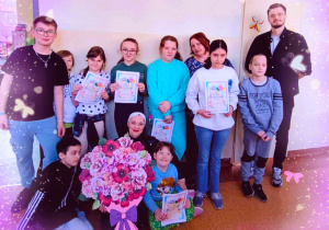 Uczniowie oraz nauczyciele ze świetlicy szkolnej pozują do wspólnego zdjęcia z wielkim papierowym bukietem kwiatów. Dziewczyny mają w rękach prezenty z okazji "Dnia Kobiet" które dostały od chłopców.