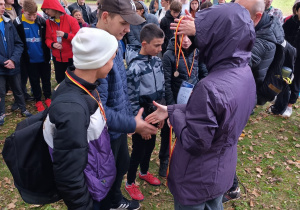 Grupa osób zgromadzona na świeżym powietrzu w parku. Dwie osoby w centrum grupy wręczają medale, podczas gdy inni obserwują. Uczestnicy mają na sobie kurtki i czapki.