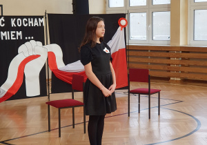 Uczennica w czarnej sukience z kotylionem stoi przed dekoracją z hasłem "Wolność kocham i rozumiem" oraz biało-czerwoną flagą.