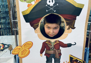 Chłopiec pozuje z głową w otworze planszy z rysunkiem pirata, tworząc zabawną scenkę zdjęciową.