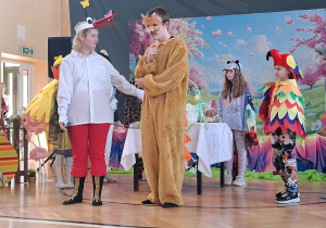 Dzieci w kolorowych kostiumach zwierząt (m.in. bocian, lis, papuga) odgrywają scenkę na szkolnej scenie z wiosenną dekoracją. Uczennica przebrana jest za bociana, a uczeń za lisa.