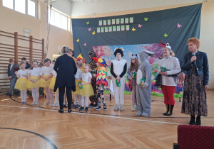 Uczniowie ustawieni w rzędzie na szkolnej sali gimnastycznej podczas zakończenia przedstawienia; pani dyrektor wręcza upominki.