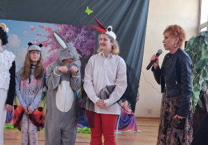 Pani dyrektor z mikrofonem przemawia obok dzieci w kostiumach zwierząt po zakończeniu przedstawienia.